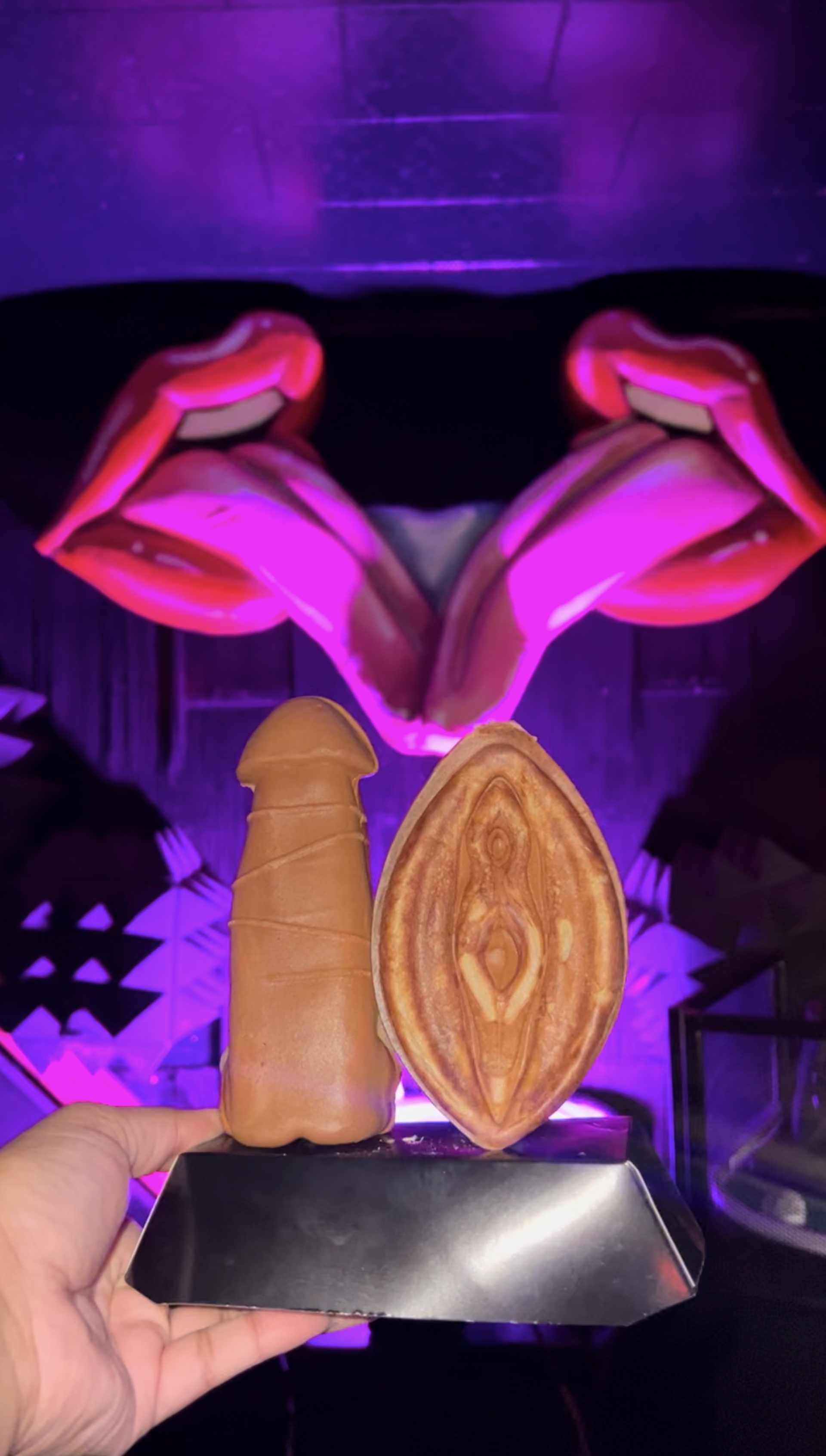 Hand holding two brown novelty sculptures — one phallic and one vulva-shaped — on a black display, set against a neon pink lips mural and purple-lit indoor art space.