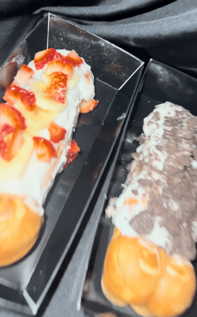 Close-up of two cream-filled éclairs on black trays — one topped with sliced strawberries and white glaze, the other with vanilla cream and chocolate shavings.