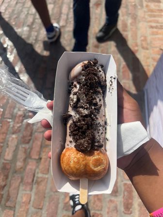Bubble-waffle ice cream on a stick topped with cookies-and-cream crumbs in a paper tray, a portable street-food dessert over brick pavement.