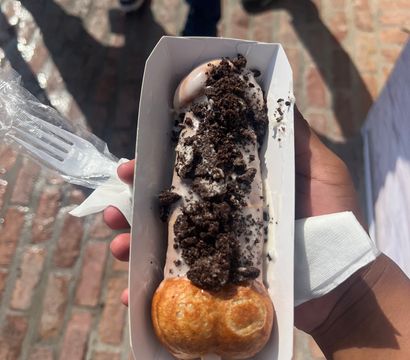 Bubble-waffle ice cream on a stick topped with cookies-and-cream crumbs in a paper tray, a portable street-food dessert over brick pavement.