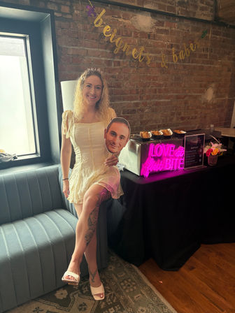 Smiling woman in a cream mini dress and tiara holding a face cutout, posing by a neon "Love at First Bite" waffle station against an exposed brick wall — festive bachelorette brunch vibe.