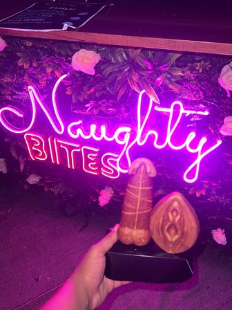 Playful scene: hand holding two adult-themed novelty pastries—phallic and vulva-shaped—on a black tray in front of a pink neon sign and faux floral wall.