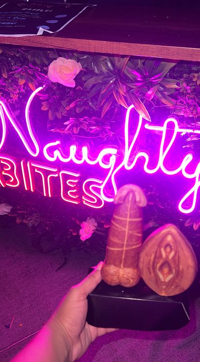 Playful scene: hand holding two adult-themed novelty pastries—phallic and vulva-shaped—on a black tray in front of a pink neon sign and faux floral wall.