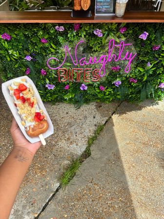 Hand holding a dessert on a stick — fried pastry topped with whipped cream, fresh strawberries and crunchy cereal — in front of a neon pink and red sign mounted on a green floral wall at an outdoor vendor.