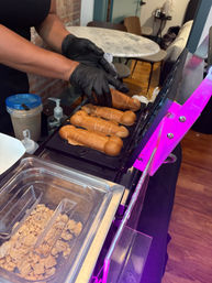 Gloved hands removing freshly baked novelty penis-shaped waffle pastries from a hot press at an indoor dessert station, with topping container and round marble table in the background.