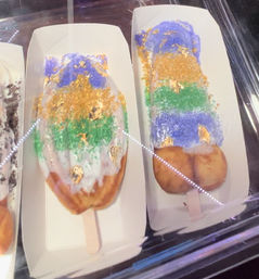 Glazed doughnut pops on wooden sticks in a bakery display case, topped with white icing, purple, green and gold sprinkles and edible gold leaf—Mardi Gras–style colors.