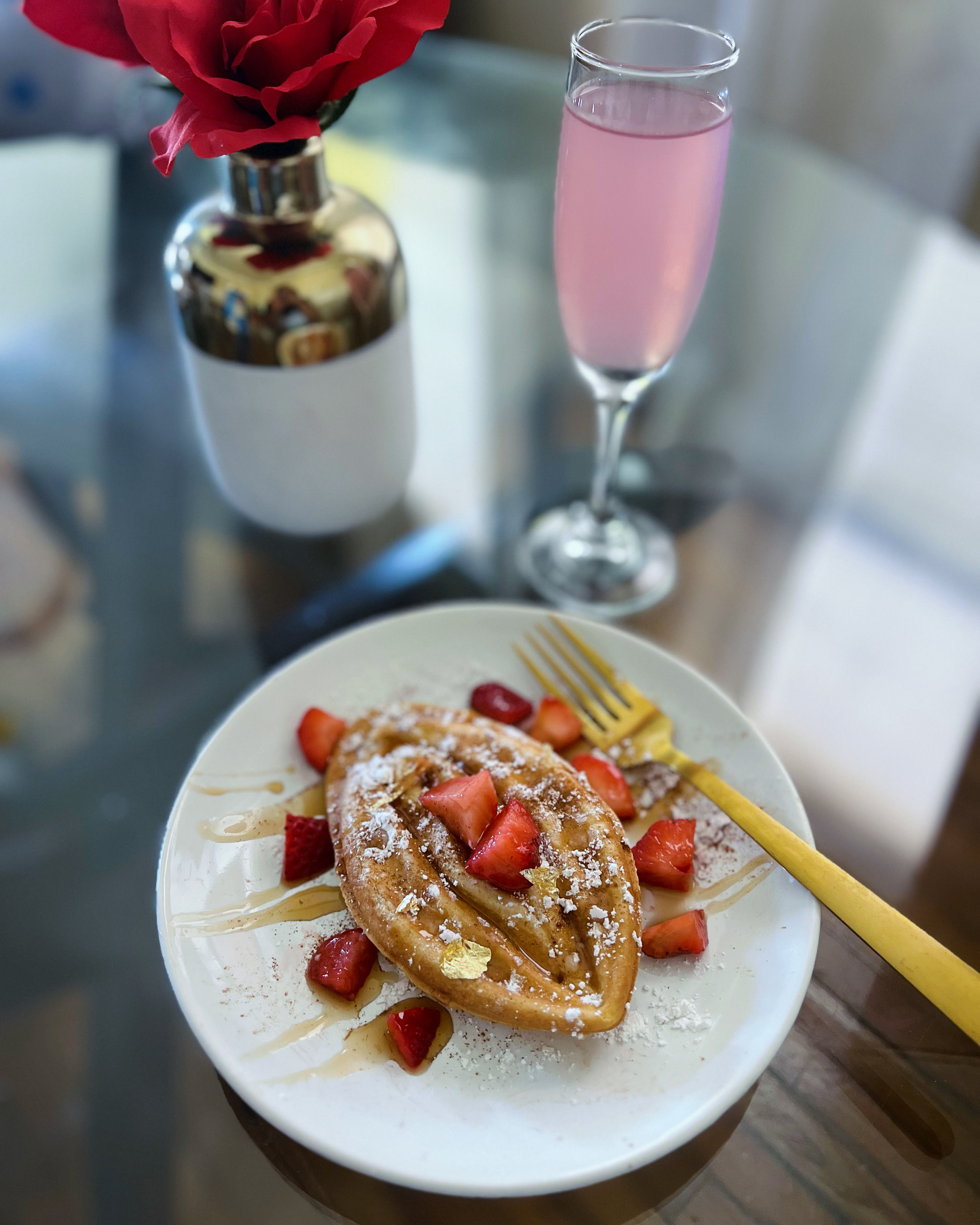 Elegant brunch scene: golden boat-shaped waffle on a white plate topped with powdered sugar, strawberries, honey drizzle and gold leaf, a gold fork, pink sparkling drink and red rose on a glass table.