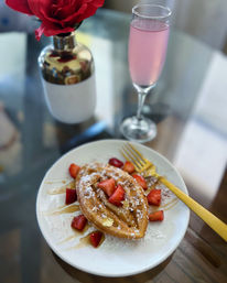 Elegant brunch scene: golden boat-shaped waffle on a white plate topped with powdered sugar, strawberries, honey drizzle and gold leaf, a gold fork, pink sparkling drink and red rose on a glass table.