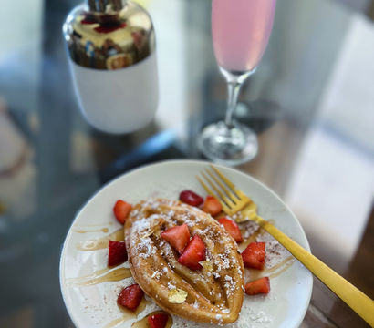 Elegant brunch scene: golden boat-shaped waffle on a white plate topped with powdered sugar, strawberries, honey drizzle and gold leaf, a gold fork, pink sparkling drink and red rose on a glass table.