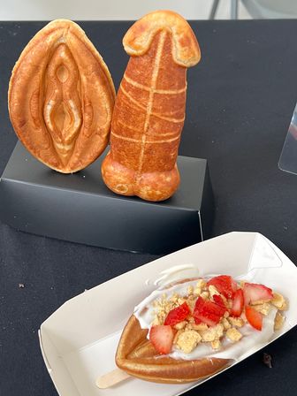 Playful food market dessert display: vulva-shaped waffle and phallic pastry on a stand, with a strawberry-and-cream waffle pop in a tray.