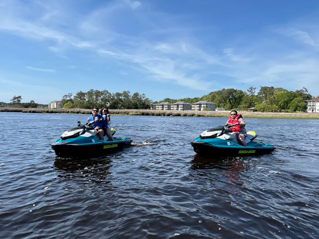 Two riders on teal jet skis cruising calm coastal bay waters near a marshy shoreline with waterfront condos under a sunny blue sky.