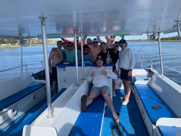 Group of friends laughing and posing on a covered pontoon boat with blue benches, one man reclining with thumbs-up, cruising a calm coastal waterway on a sunny day.