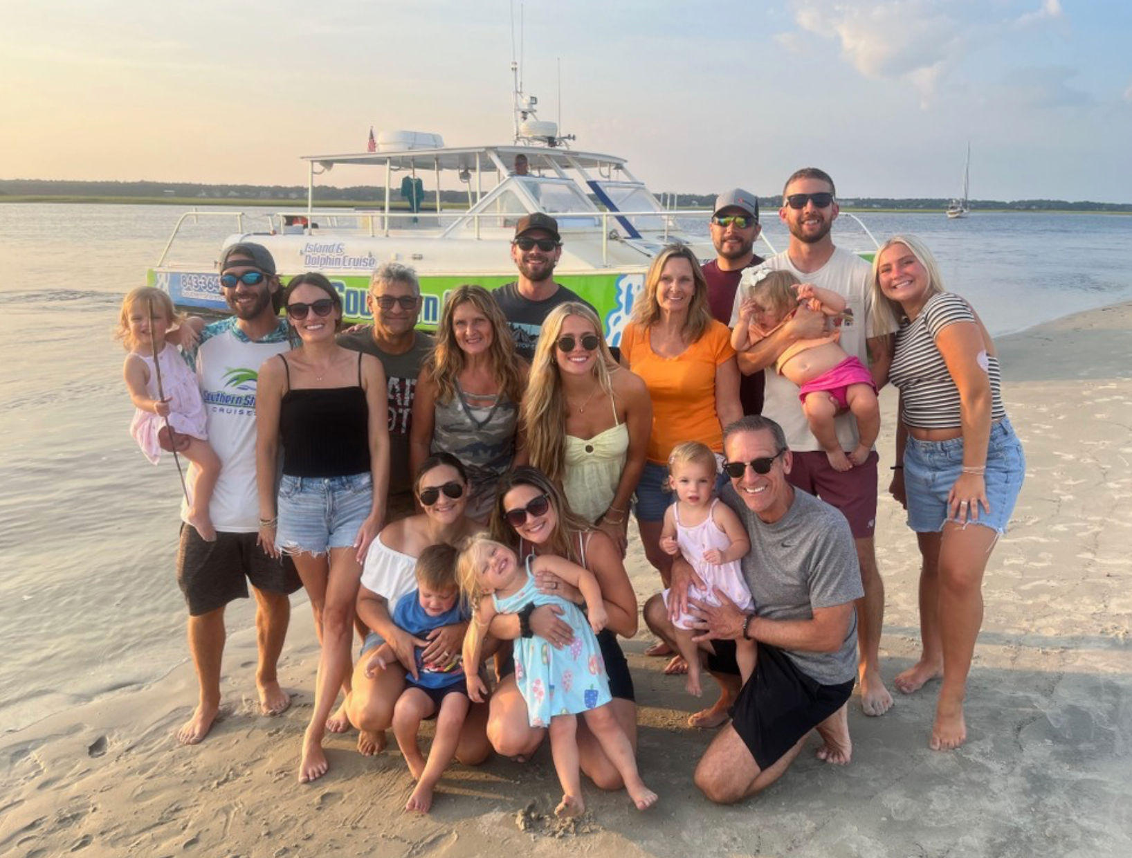 Multigenerational group of family and friends posing on a sandy beach at sunset, children in front and a white boat anchored on calm coastal water behind them.