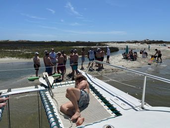 Sunny coastal sandbar scene with people wading and socializing in shallow water beside a beached catamaran; one person lounges on the boat’s trampoline, with beach chairs, umbrella, dunes and salt marsh grasses under a clear blue sky.