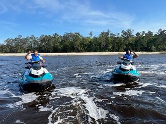 Two riders in blue life vests waving from turquoise jet skis on a calm coastal river near a sandy beach and tree-lined shoreline under a bright blue sky