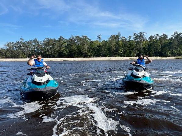 Two riders in blue life vests waving from turquoise jet skis on a calm coastal river near a sandy beach and tree-lined shoreline under a bright blue sky