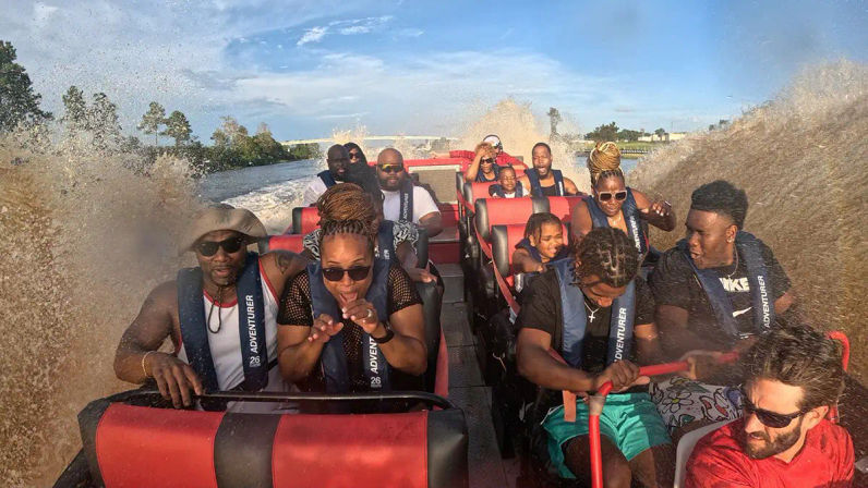 Group of passengers in life jackets on a red jet boat, reacting and laughing as a giant splash soaks them on a sunny river thrill ride.