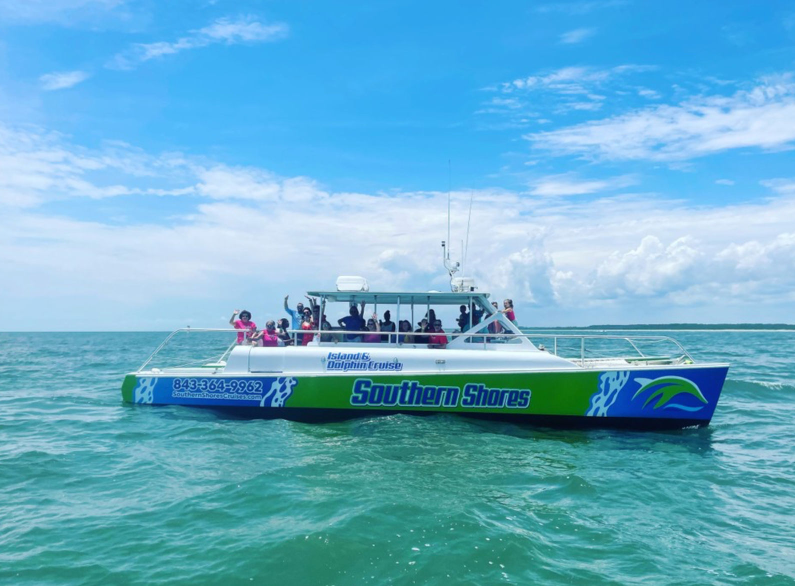 Colorful green-and-blue passenger catamaran dolphin-watch tour with people waving on turquoise coastal waters under a sunny blue sky with scattered clouds.