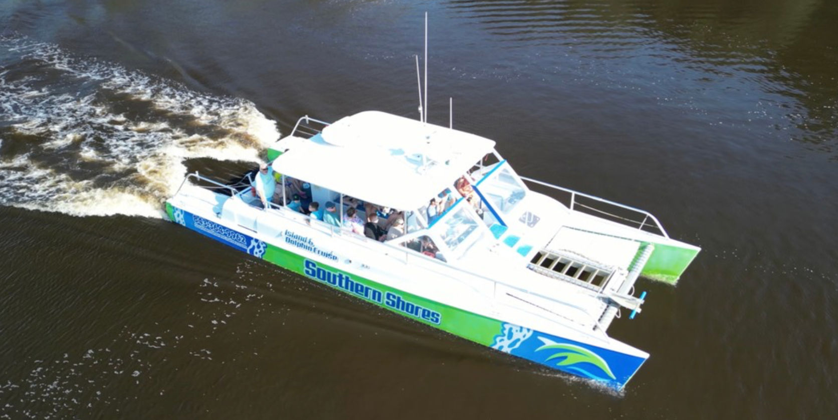 Aerial view of a green-and-blue catamaran tour boat with passengers cruising on a brown river, leaving a foamy white wake