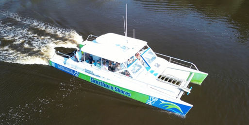 Aerial view of a green-and-blue catamaran tour boat with passengers cruising on a brown river, leaving a foamy white wake