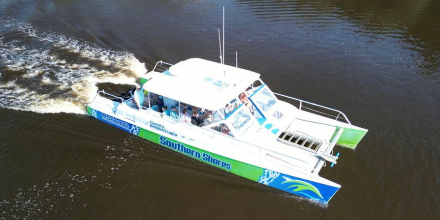 Aerial view of a green-and-blue catamaran tour boat with passengers cruising on a brown river, leaving a foamy white wake