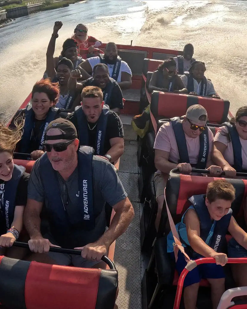 Passengers wearing life jackets on a red-and-black jet boat speeding down a sunlit river, gripping safety bars and cheering as water spray and the boat’s wake trail behind.