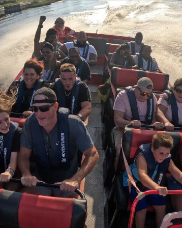 Passengers wearing life jackets on a red-and-black jet boat speeding down a sunlit river, gripping safety bars and cheering as water spray and the boat’s wake trail behind.