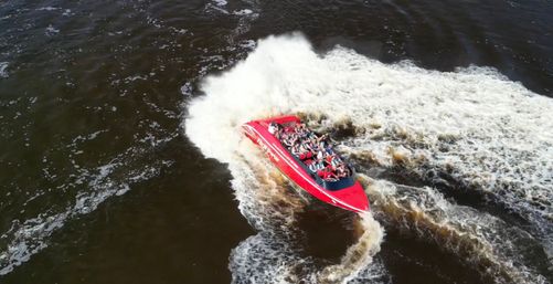 Red jet boat with passengers on a river tour making a sharp turn, throwing a dramatic white spray and swirling wake across dark water