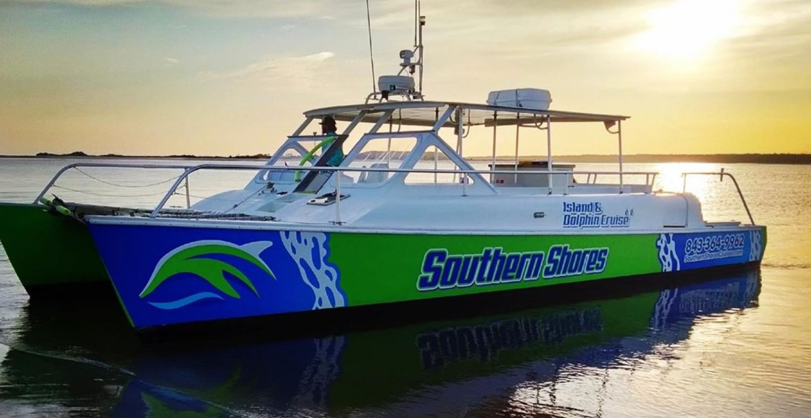 Bright green-and-blue catamaran tour boat with open deck and canopy on calm coastal waters at sunset, ready for a dolphin-watching cruise.