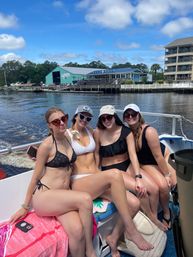 Four women in swimsuits and sunglasses smiling and posing on a motorboat under a bright blue sky, with a calm river, marina docks and waterfront restaurants in the background — relaxed summertime boating scene.
