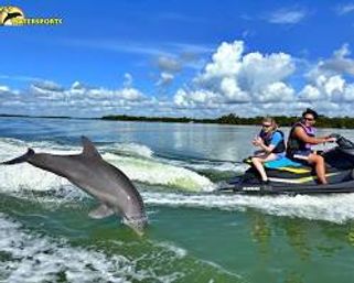 Playful dolphin leaping through a wake beside a jet ski with two riders on calm green coastal waters under a bright blue sky with puffy clouds