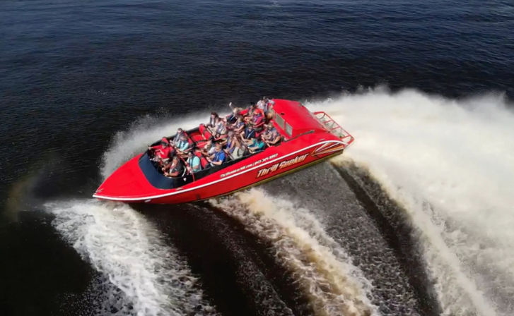 Red high-speed tour boat loaded with passengers in life jackets carving a sharp turn and spraying a dramatic white wake across dark river water