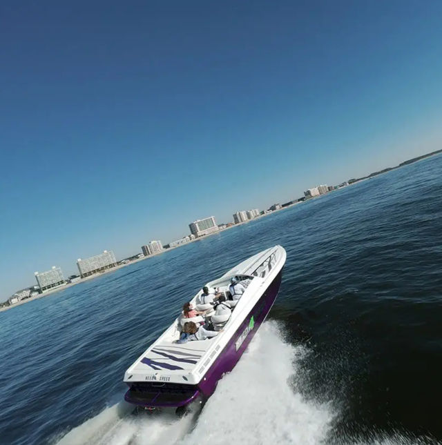 Purple-and-white speedboat slicing across calm ocean water near a coastline lined with beachfront high-rise condos under a clear blue sky, passengers enjoying the ride and a white wake trailing behind.