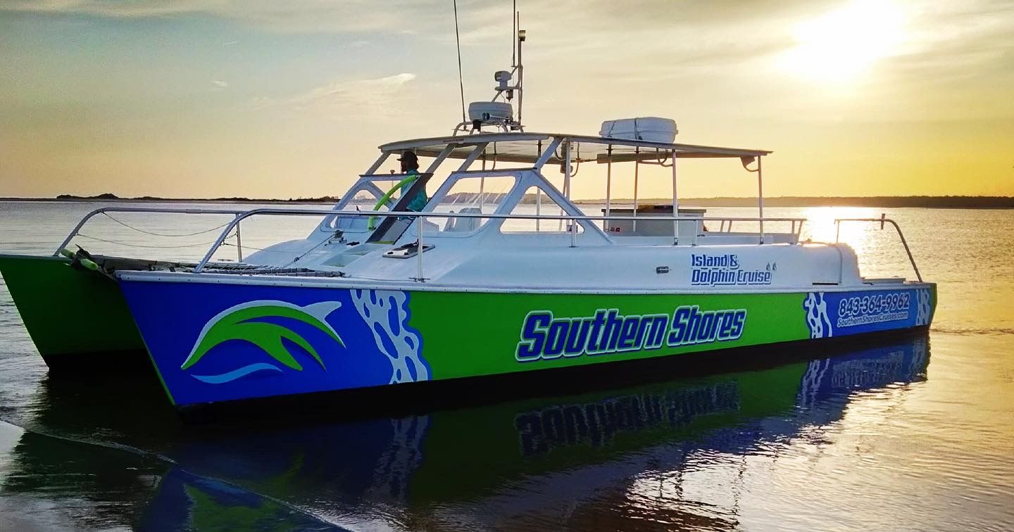 Colorful green-and-blue catamaran dolphin tour boat with open deck reflected on calm coastal waters at sunset.