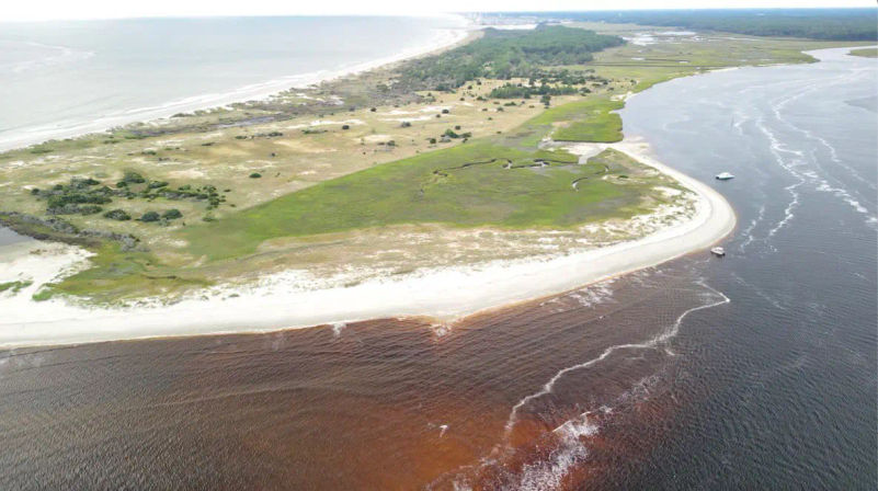 Aerial view of a curved sandy barrier island and green salt marsh, white beach with two boats anchored offshore, and brown estuary water swirling where it meets the open ocean.