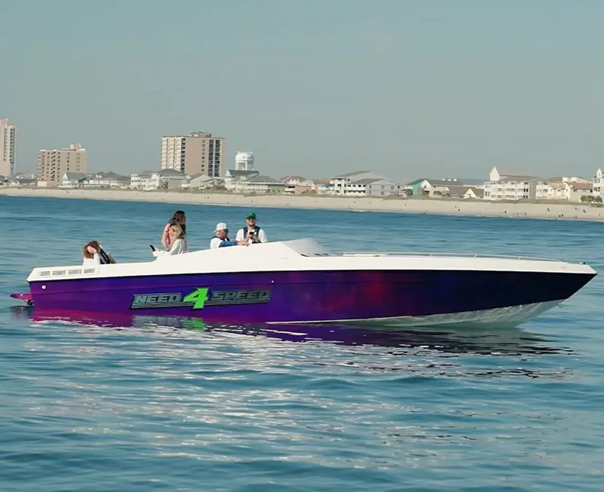 Purple speedboat with bright green "NEED 4 SPEED" decal and passengers cruising calm blue water near a sandy beachfront and low-rise coastal buildings