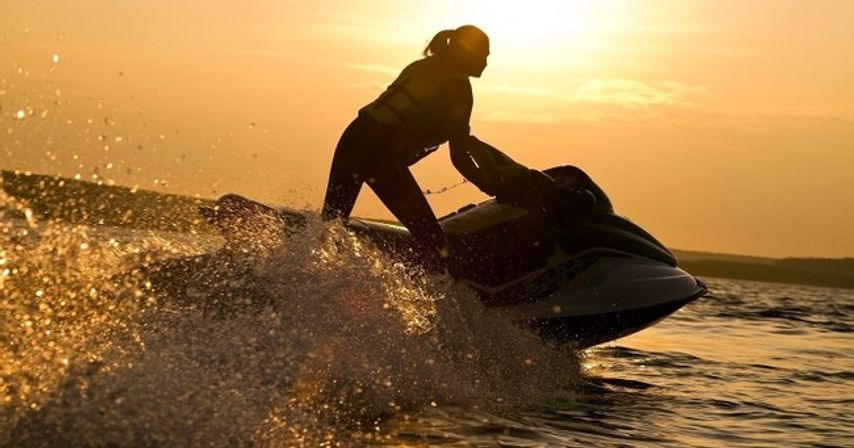 Silhouette of a person in a life jacket riding a jet ski at sunset, golden spray and waves against a coastal horizon — energetic watersports scene.