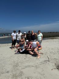 Group of friends posing on a sandy beach sandbar beside a calm coastal inlet and marsh, wearing summer beachwear on a sunny day under a clear blue sky.