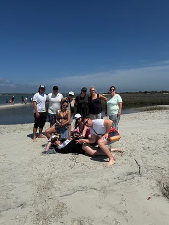 Group of friends posing on a sandy beach sandbar beside a calm coastal inlet and marsh, wearing summer beachwear on a sunny day under a clear blue sky.