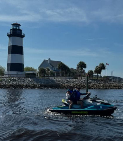 Two riders on a teal jet ski cruising across coastal water near a black-and-white striped lighthouse on a rocky shoreline with palm trees and blue sky