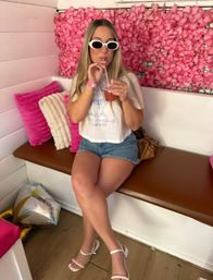 Person in white sunglasses and denim shorts sipping a pink cocktail while seated on a bench against a vibrant pink flower wall in a trendy pink-themed lounge