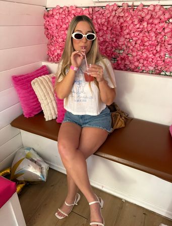 Person in white sunglasses and denim shorts sipping a pink cocktail while seated on a bench against a vibrant pink flower wall in a trendy pink-themed lounge