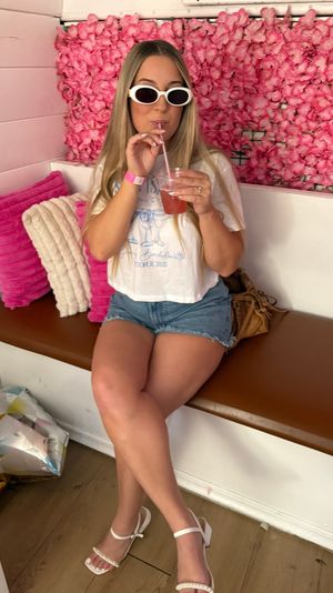 Person in white sunglasses and denim shorts sipping a pink cocktail while seated on a bench against a vibrant pink flower wall in a trendy pink-themed lounge