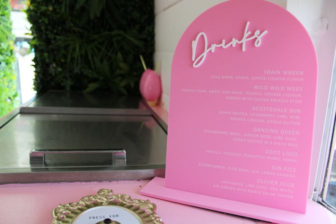 Playful pastel pink arched drinks menu listing cocktails on a counter beside a stainless steel service hatch and lush green wall — bright, modern cocktail bar setup.