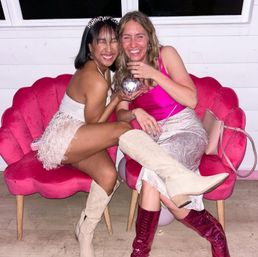 Two smiling friends at an indoor pink-themed birthday party seated on scallop velvet chairs, one wearing a birthday tiara and white fringe skirt, the other in a fuchsia top and patterned midi skirt, both wearing knee-high boots and sharing a small disco ball prop.