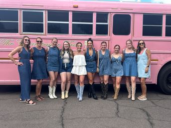 Ten smiling women in denim dresses, rompers and a white outfit, many wearing cowboy boots, posing together in front of a bright pink party bus on a sunlit pavement.
