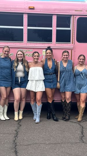 Ten smiling women in denim dresses, rompers and a white outfit, many wearing cowboy boots, posing together in front of a bright pink party bus on a sunlit pavement.