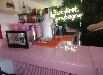 Stylish pink cocktail bar counter with bottles, a champagne bottle, tip jar and pink straws under a glowing neon sign that reads 'But first, champagne' against a leafy green backdrop.