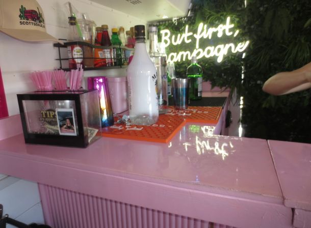 Stylish pink cocktail bar counter with bottles, a champagne bottle, tip jar and pink straws under a glowing neon sign that reads 'But first, champagne' against a leafy green backdrop.