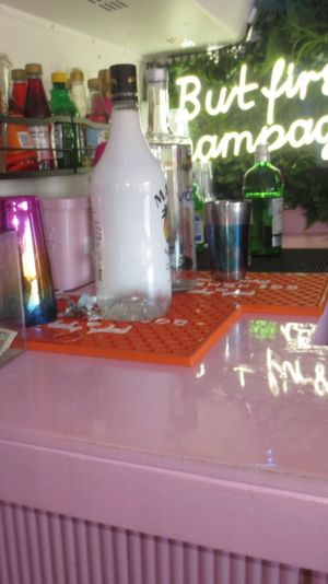 Stylish pink cocktail bar counter with bottles, a champagne bottle, tip jar and pink straws under a glowing neon sign that reads 'But first, champagne' against a leafy green backdrop.